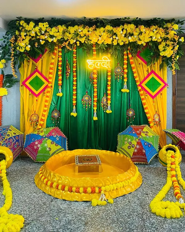 This is a complete Haldi ceremony setup designed for an intimate home function. It features a traditional low seating arrangement, a backdrop of bright yellow and green drapes, and is topped with lush marigold garlands. A custom Hindi 'Haldi' neon sign adds a modern touch, complemented by colorful Rajasthani umbrellas.