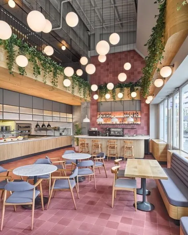 We designed this spacious, double-height cafe to feel vibrant and full of life. The design features a bold pink tiled floor, an array of spherical pendant lights for dramatic effect, and hanging greenery to soften the industrial architecture, creating an energetic and welcoming atmosphere for customers.