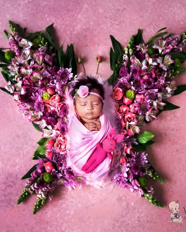 A newborn baby girl wrapped in pink, nestled amongst a beautiful arrangement of fresh flowers forming butterfly wings. This artistic newborn portrait is a perfect way to celebrate your new arrival.