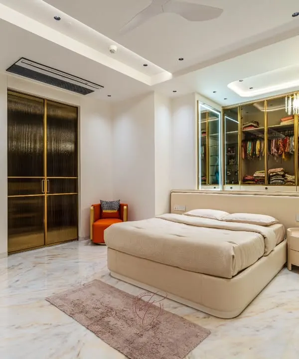 A wide view of a master bedroom that exudes luxury and tranquility. The design features a stunning walk-in closet with textured glass and gold-finished doors, which serves as a beautiful backdrop to the plush, neutral-toned bed.