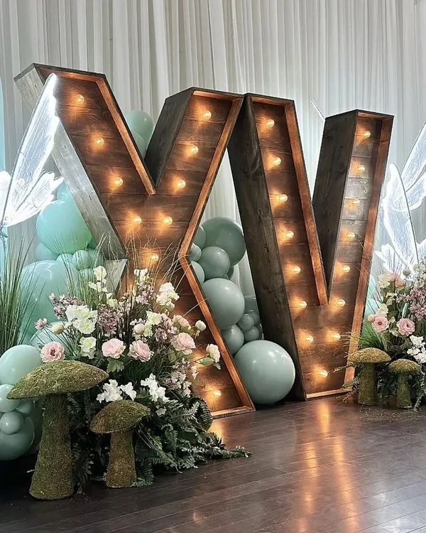 An enchanted forest themed decor featuring large rustic marquee letters. This whimsical setup includes pastel balloons, illuminated butterfly figures, and mossy mushroom props.
