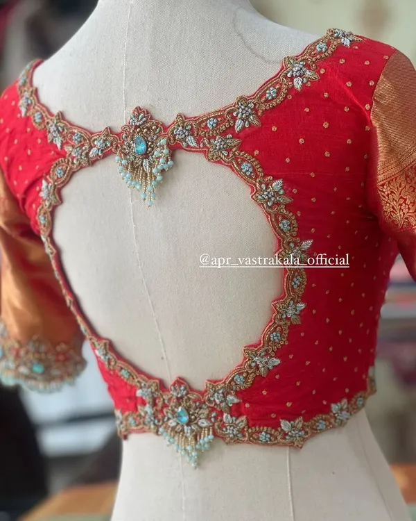 A bold statement back design. This bright red bridal blouse features a dramatic heart-shaped cutout, beautifully framed with intricate floral zardosi and light blue stone work.