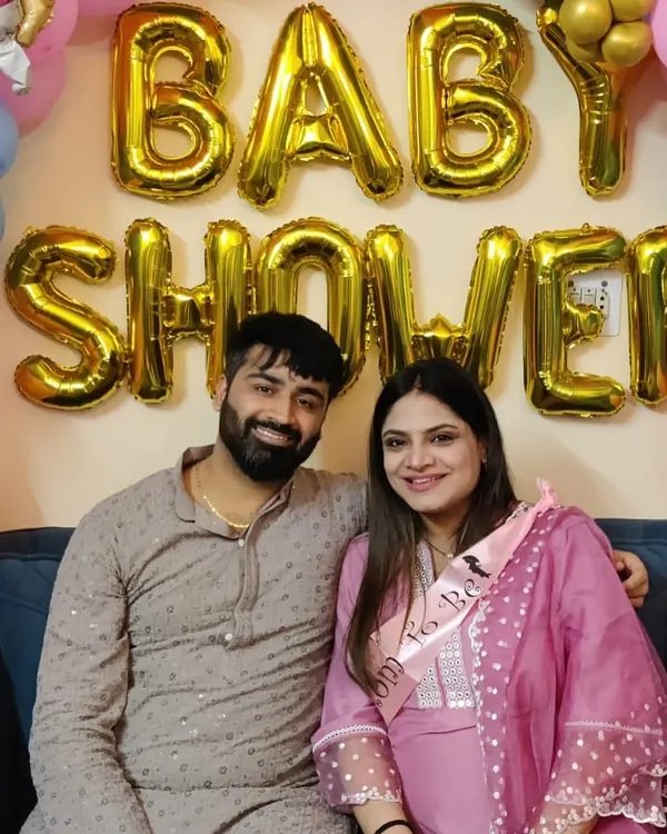A happy couple posing in front of a simple 'Baby Shower' decoration featuring large gold foil letters.