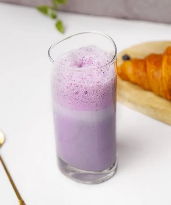 A bright blueberry smoothie in a tall glass, paired with a croissant in a clean, lifestyle cafe setting.