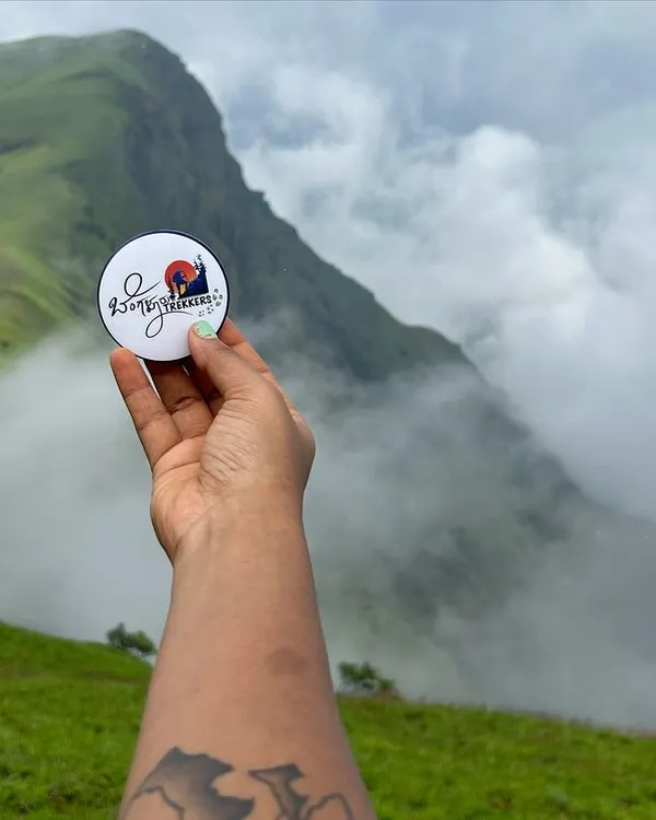 Our brand badge held up against the stunning, cloud-covered peaks of the Kudremukha range. This is what monsoon trekking in Karnataka is all about.