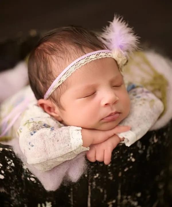 A classic newborn pose, with the baby sleeping peacefully in a dark bucket, wearing a delicate floral outfit and a lace headband.