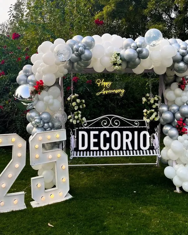 This is the full view of the 25th-anniversary swing setup in a garden. The combination of the balloon arch, neon sign, and large marquee numbers creates a stunning photo opportunity for the celebrating couple and their guests.