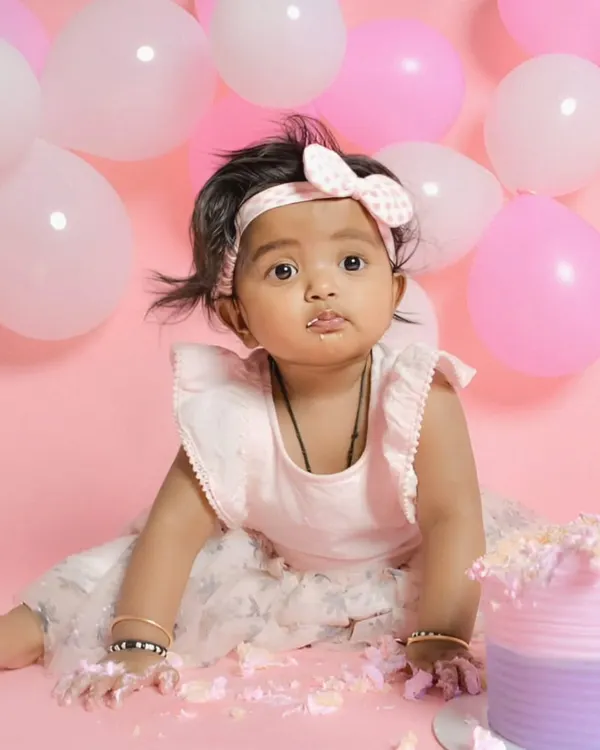 A classic cake smash moment. A cute baby with frosting on her face looks at the camera amidst a fun pink balloon backdrop, perfectly capturing the messy joy of a first birthday.