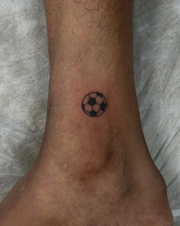 For the love of the game. A tiny, minimalist football tattoo on the ankle, created with clean lines and solid black shading. It's a small but powerful symbol of passion.