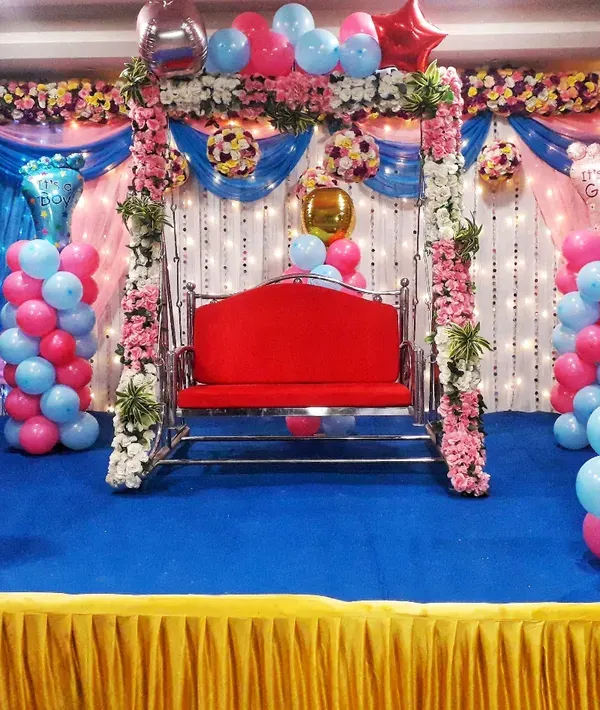 A fun and colorful baby shower stage for a baby girl. The setup includes pink and blue balloon pillars with baby-shaped foil balloons, a decorated swing, and a floral backdrop.