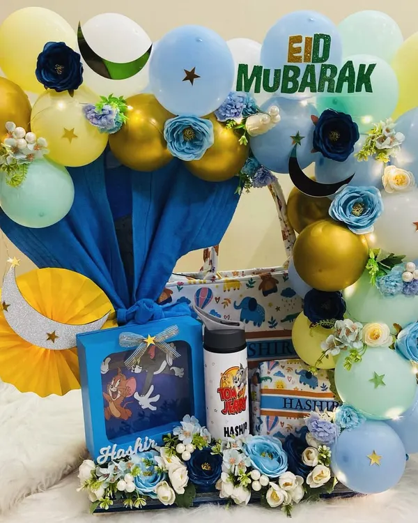 An elegant Eid Mubarak hamper for a little one. This stunning arrangement features a blue and gold theme with balloons, flowers, and personalised gifts like a Tom & Jerry t-shirt and a custom water bottle for 'Hashir'.
