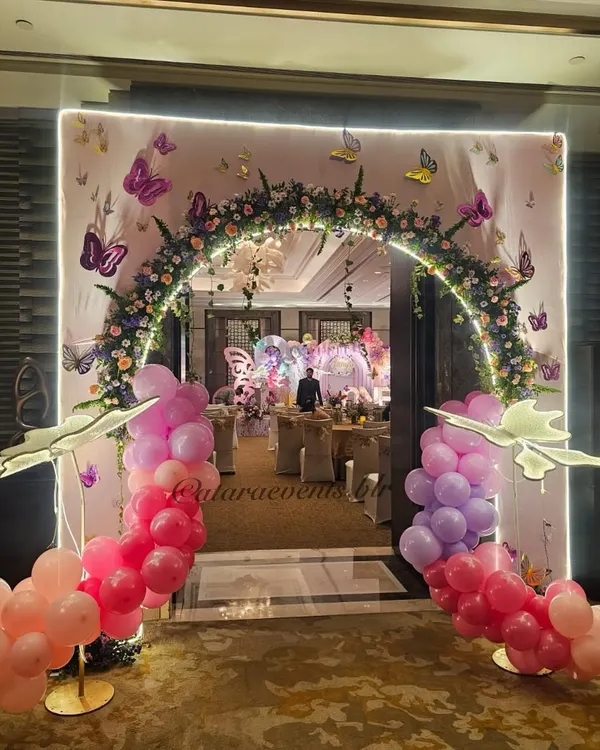 Welcome to a magical wonderland. This stunning entrance features a floral arch with butterfly accents, illuminated by soft lights and framed by pink and purple balloon garlands.