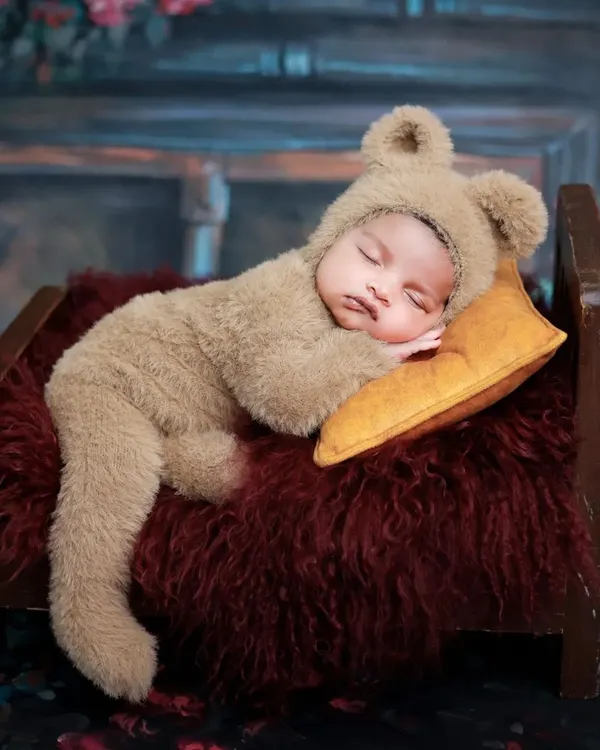 A baby dressed in a fuzzy bear costume sleeps peacefully on a miniature wooden bed. This is one of our most requested adorable prop setups.