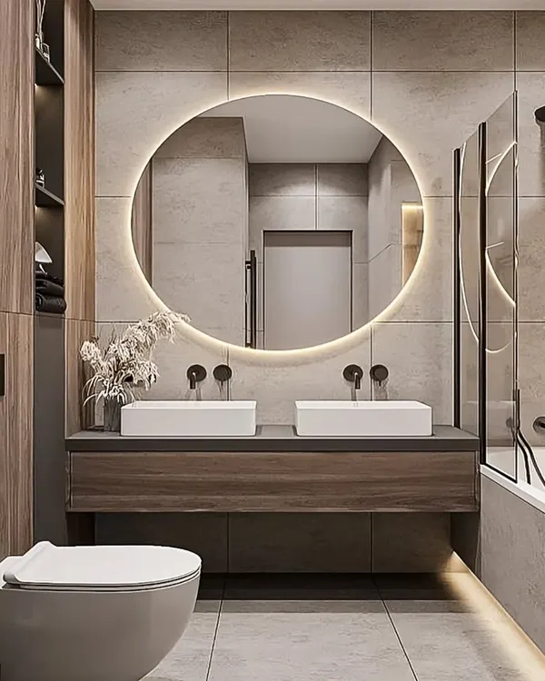 A modern bathroom featuring a floating wood-finish vanity with dual sinks. The large, round backlit mirror and under-vanity lighting create a warm and inviting ambiance.