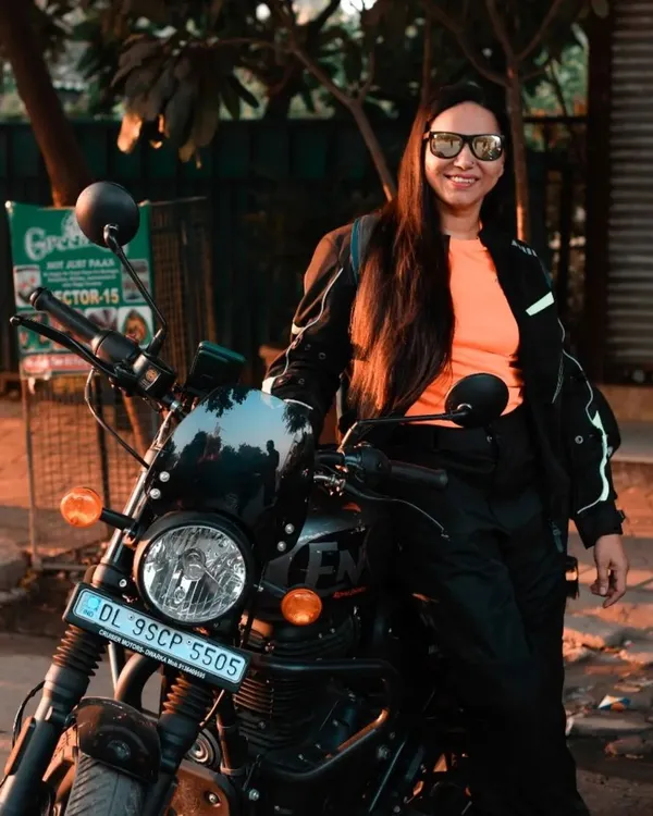 A confident female rider posing with her Royal Enfield Hunter. The warm evening light and urban setting create a cool, atmospheric portrait that captures the essence of modern motorcycling culture.