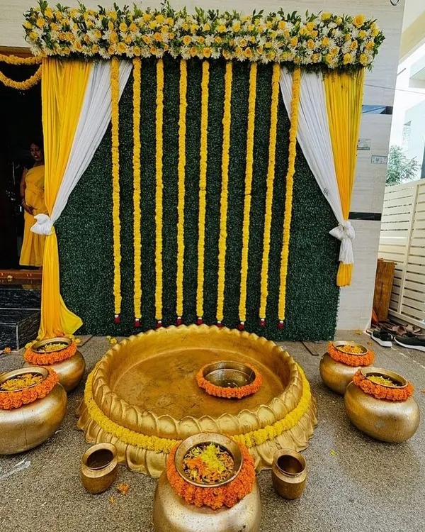 A traditional Haldi setup featuring a central brass urli for the ceremony, surrounded by smaller pots. The backdrop combines a lush green wall with vertical strings of marigold flowers and elegant yellow and white drapes.