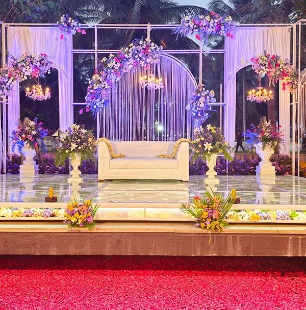 A panoramic view of a glamorous outdoor reception stage. The design features elegant white arches, cascading string lights, and lush floral arrangements, with a pathway of red petals leading to the stage.