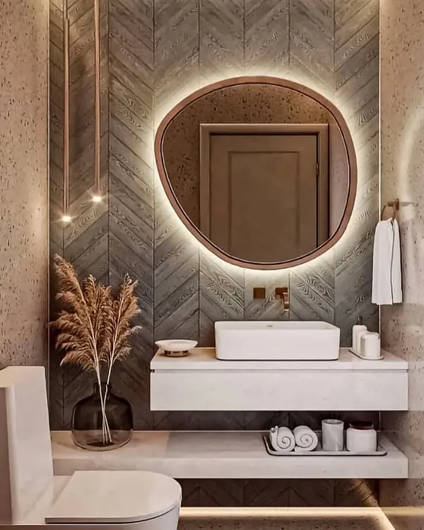 A stylish powder room featuring a floating white vanity and a uniquely shaped backlit mirror. The chevron-patterned tile wall adds texture and warmth to the space.