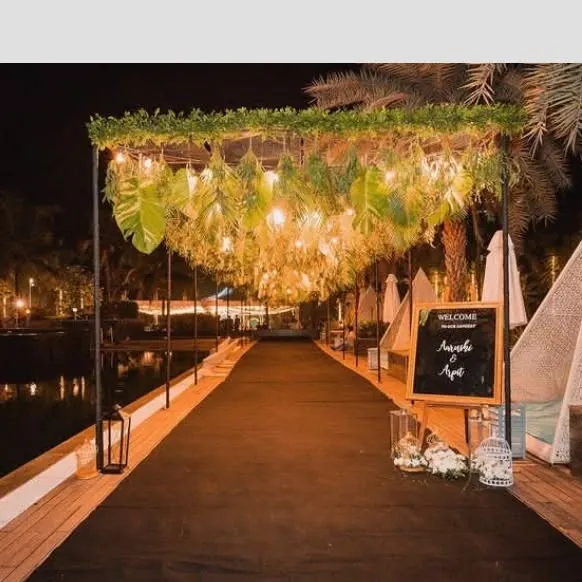 A stunning walkway for an elegant outdoor event by the water. The entrance features a canopy decorated with lush green leaves and hanging lights, creating a magical pathway for guests.