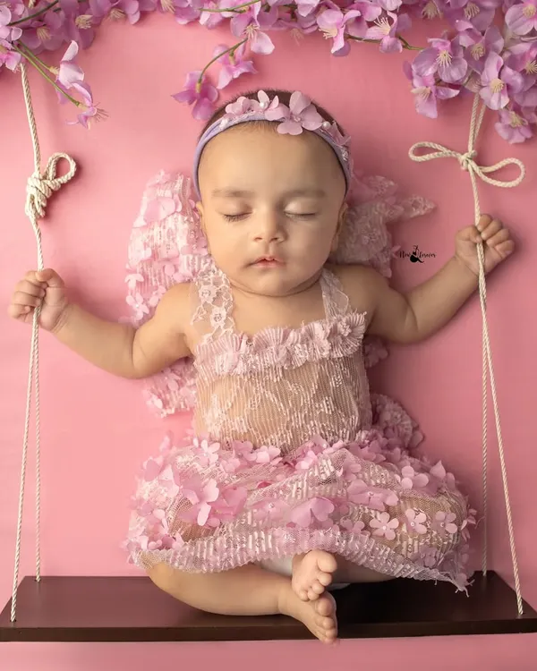 A little angel sleeping peacefully on a swing. This setup uses delicate props like the floral wings and a soft pink backdrop to create a dreamy, ethereal portrait.