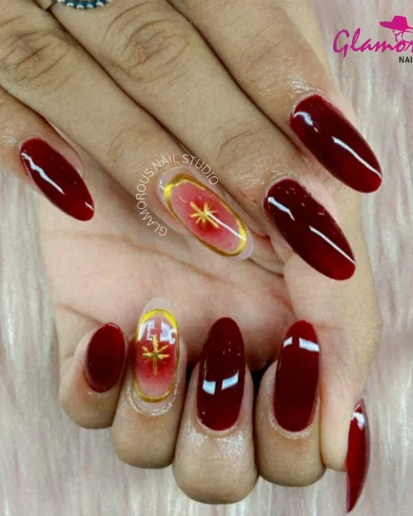 A look of pure luxury and drama. This design features long, almond-shaped nails in a deep, glossy red, with accent nails showcasing a celestial gold starburst on a sheer base.