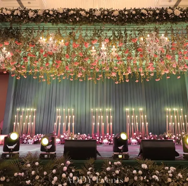 A Sangeet stage that exudes pure splendor. This design features a ceiling dripping with flowers and chandeliers, a teal backdrop, and candle-lit accents, creating a luxurious and dramatic setting for performances.