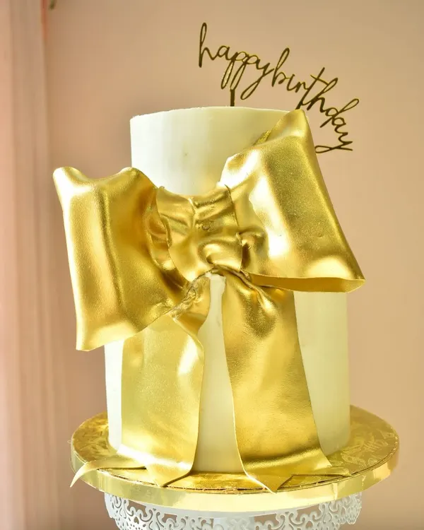 A statement of pure elegance. This two-tier white cake is wrapped in a large, luxurious, hand-sculpted golden bow, making it a glamorous centerpiece for a sophisticated birthday or anniversary celebration.