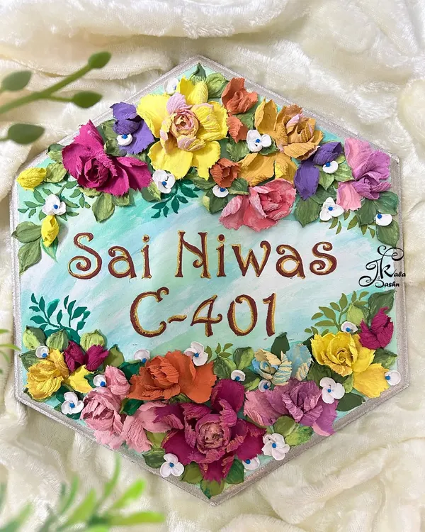 A beautiful hexagonal nameplate for "Sai Niwas," featuring a refreshing light blue base and a wreath of vibrant, multi-colored sculpted flowers. The brown lettering stands out beautifully against the background.