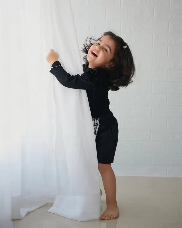 A joyful, candid shot of a little girl in a black outfit laughing as she plays with a white curtain.