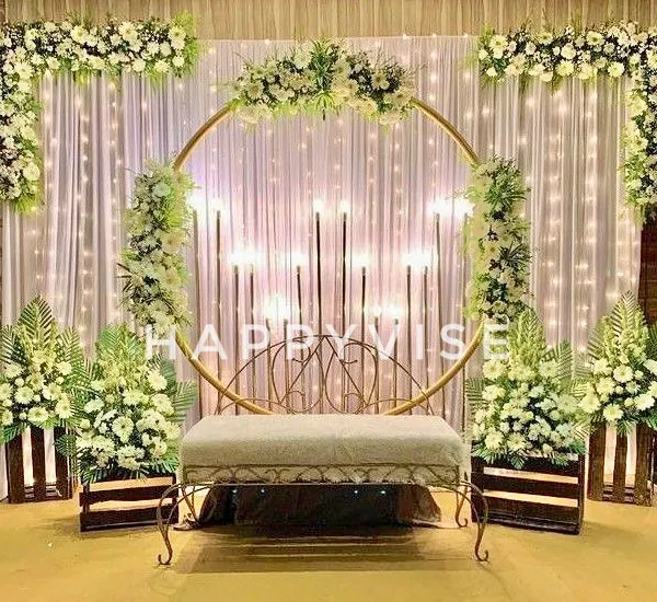 A beautiful stage setup for an engagement, featuring a golden ring backdrop adorned with white flowers and surrounded by fairy lights. The design creates a warm and magical ambiance.