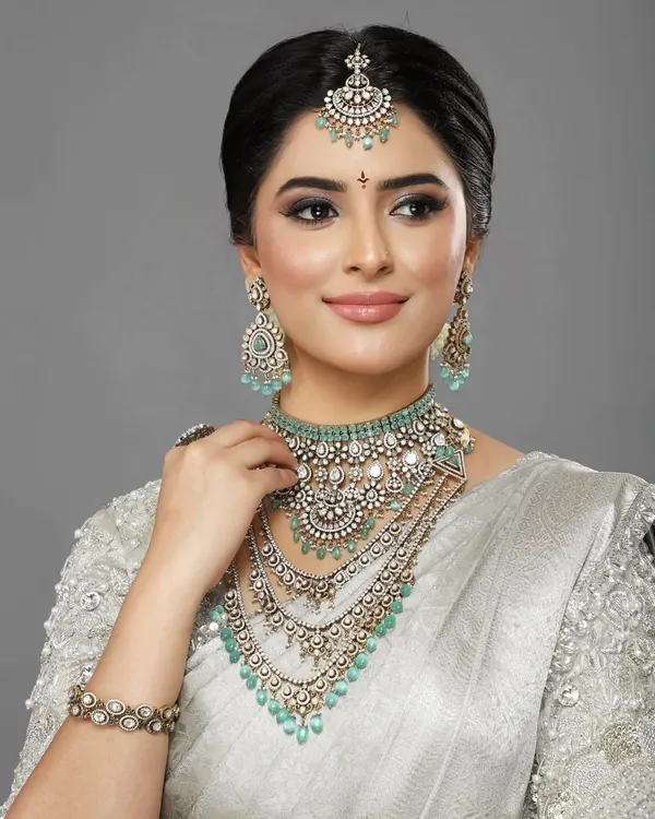 An editorial-style portrait featuring a stunning silver saree and layered diamond and mint-green jewelry. The makeup is clean and luminous, with a focus on flawless skin and a defined eye.