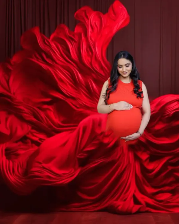A full-body portrait of a mother-to-be in a magnificent red gown. The fabric is arranged to create a sense of movement and grandeur against a matching red backdrop.