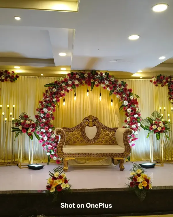 A grand reception stage decoration in Halsur. The setup features a large floral arch of red and white gerberas, a golden diwan, and rows of elegant stand-up lights on either side.