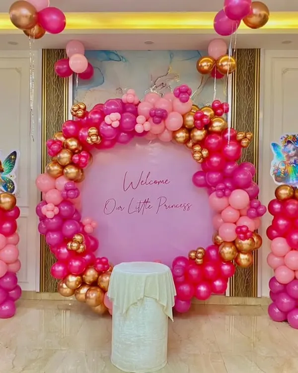 A beautiful welcome for a little princess! This elegant setup features a full ring backdrop in shades of pink and gold, with fairy cutouts and matching balloon pillars, perfect for making a grand statement in your home.