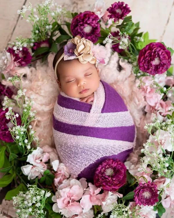 A classic and beautiful newborn portrait. The baby is wrapped snugly in purple and surrounded by a wreath of matching flowers, creating a peaceful and artistic image.
