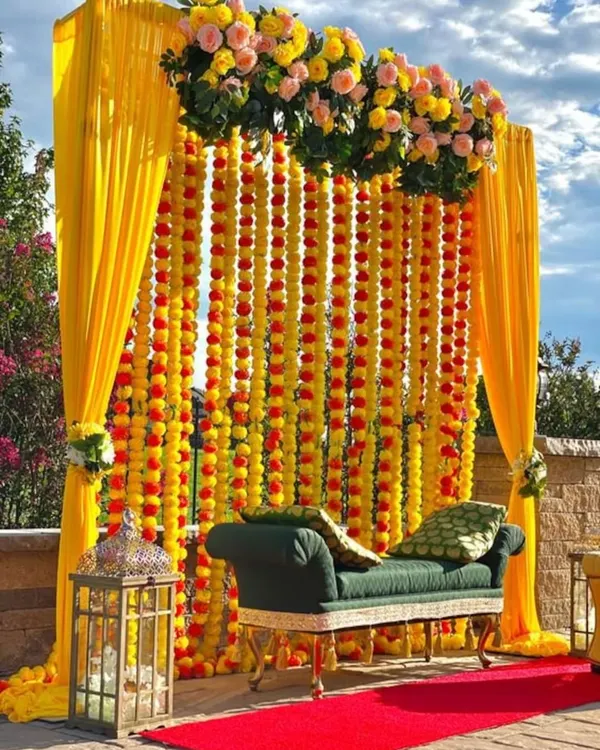 A classic and vibrant Haldi seating area we designed for an outdoor celebration. The backdrop features dense strings of yellow and orange marigolds, framed by bright yellow drapes, creating a perfect spot for photos.