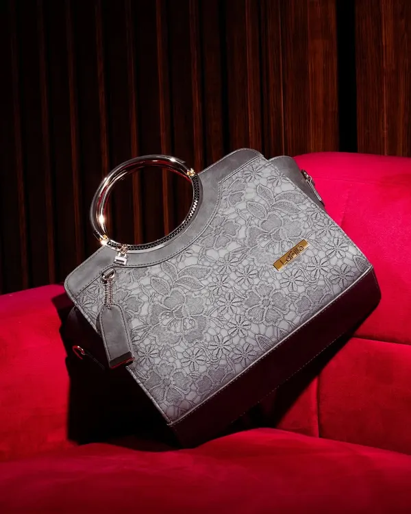 This elegant grey handbag with a unique metal ring handle is placed on a rich, red velvet couch. The luxurious setting and lighting highlight the bag's sophisticated design.