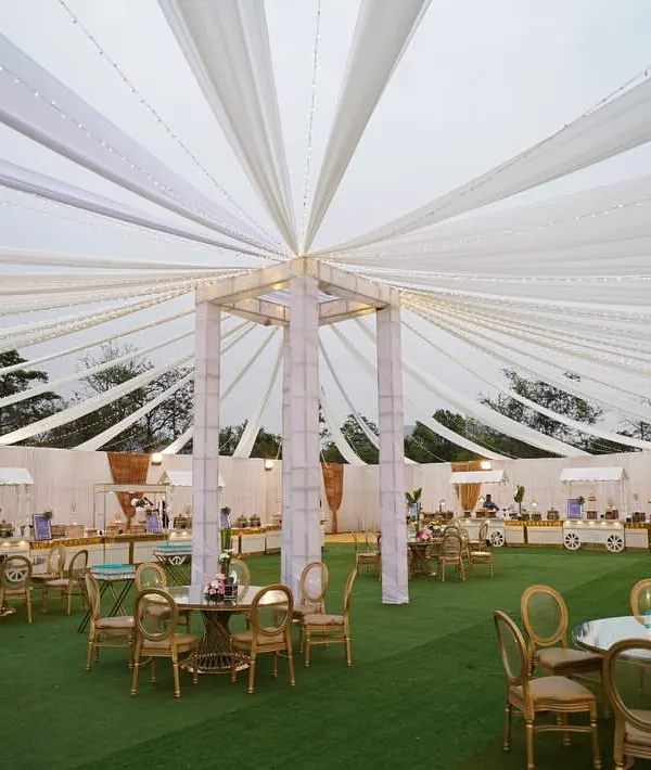 A wide-angle view of an outdoor event setup, featuring a magnificent white draped tent over lush green turf. This creates a bright and airy atmosphere for a daytime celebration.