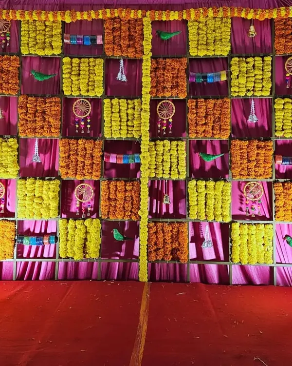 A traditional backdrop for a baby shower, featuring a checkerboard pattern of yellow and orange marigolds against a pink fabric. The small dreamcatchers and parrots add a unique, decorative touch.