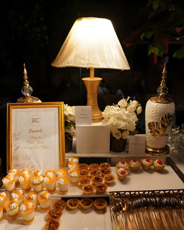 An elegant dessert table featuring a variety of sweet treats under soft lamp lighting. The display includes dessert shots, pecan tarts, and mini cheesecakes, complete with golden cutlery.