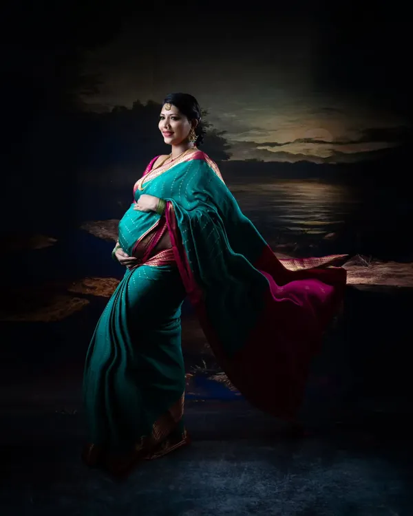 An artistic maternity portrait of a woman in a teal and pink saree, with the fabric flowing beautifully against a painted seascape backdrop.