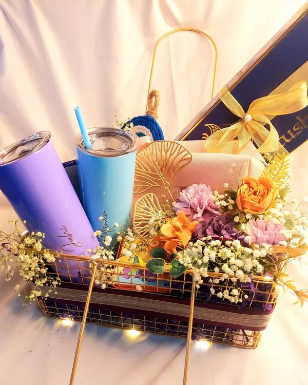 A vibrant and colorful wedding gift hamper for a fun-loving couple. This basket includes two personalized tumblers, a jewelry box, and other goodies, all surrounded by a beautiful floral arrangement.