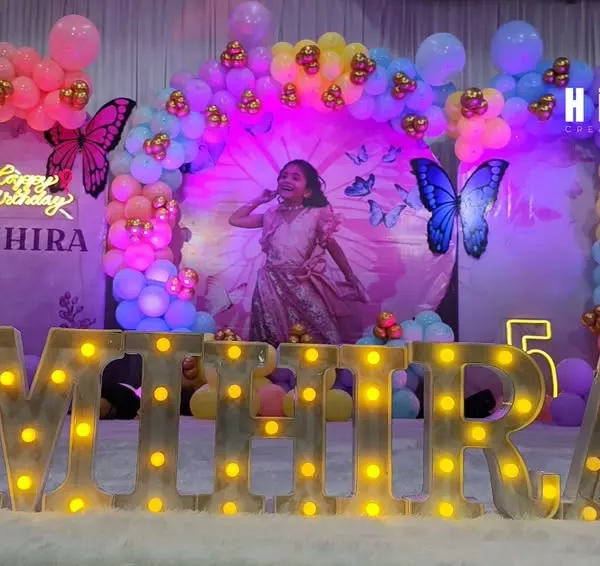 This butterfly-themed birthday party was a dream to create. I designed a magical backdrop with a photo of the birthday girl, surrounded by a rainbow balloon arch and floating butterflies, with her name "MIHIRA" lit up in large marquee letters.