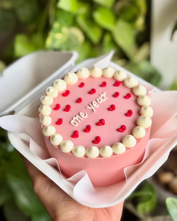 A tiny cake for a big milestone. This pink bento cake celebrates "one year" with a classic pearl border and tiny red hearts, packed and ready to gift.