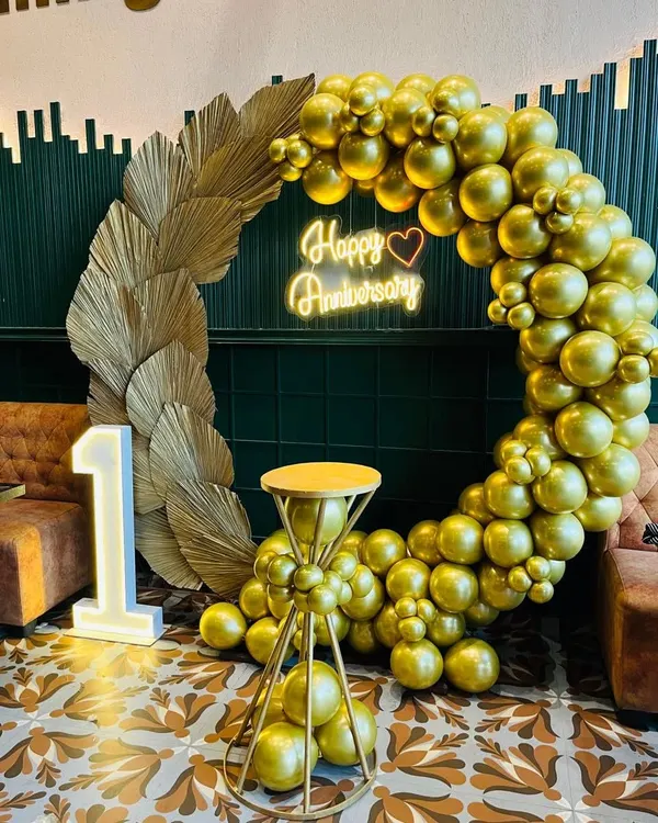An elegant and sophisticated first-anniversary decoration. This setup features a monochrome gold balloon arch, a 'Happy Anniversary' neon sign, and a large light-up marquee number '1'.