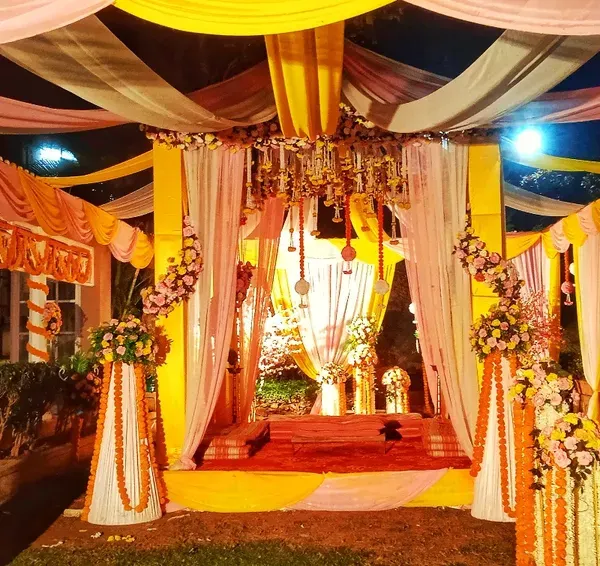 A stunning outdoor evening mandap with a ceiling of draped yellow and pink fabric, illuminated by a central chandelier and floral arrangements.