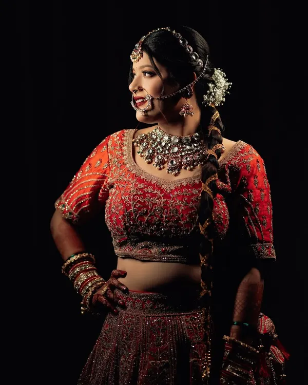 A striking bridal portrait against a dark background. The side profile and dramatic lighting highlight the intricate details of her red lehenga, jewelry, and confident pose.