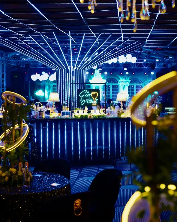 A chic and modern bar setup for a Sangeet night, featuring a "Gin O'Clock" neon sign and a facade of vertical blue LED lights that create a high-energy, club-like atmosphere.