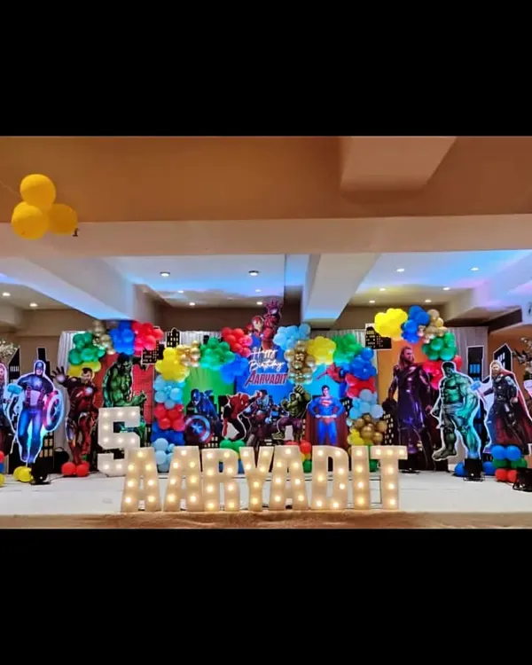 An epic superhero assembly for a 5th birthday party. The stage is packed with cutouts of famous heroes, colorful balloon clusters, and the birthday boy's name in large marquee letters, creating a powerful and exciting atmosphere.