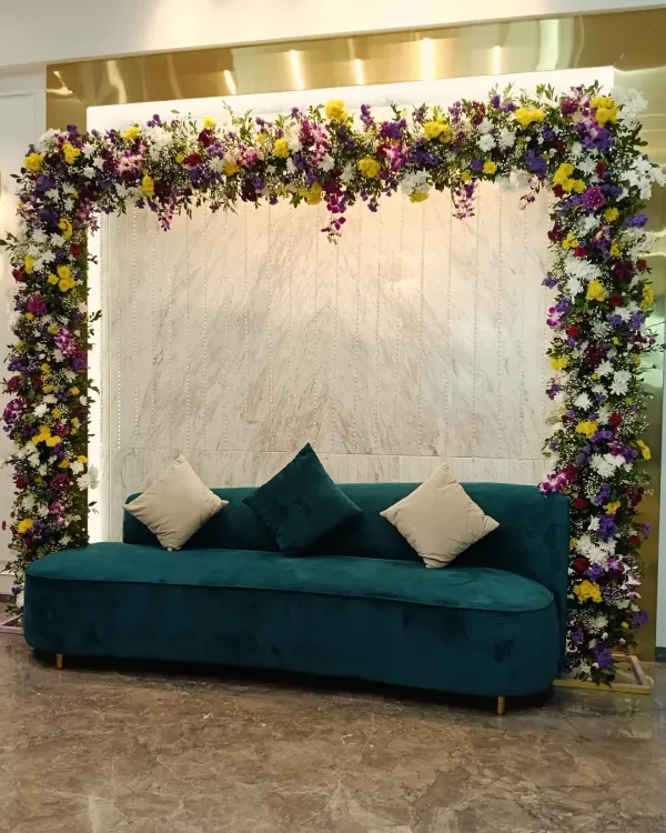 A modern and chic engagement backdrop. The rectangular arch is covered in a mix of colorful flowers, perfectly framing the stylish teal velvet sofa for photos.